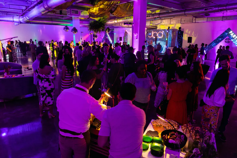 Crowded indoor party with purple lighting, people mingling around tables and cocktails