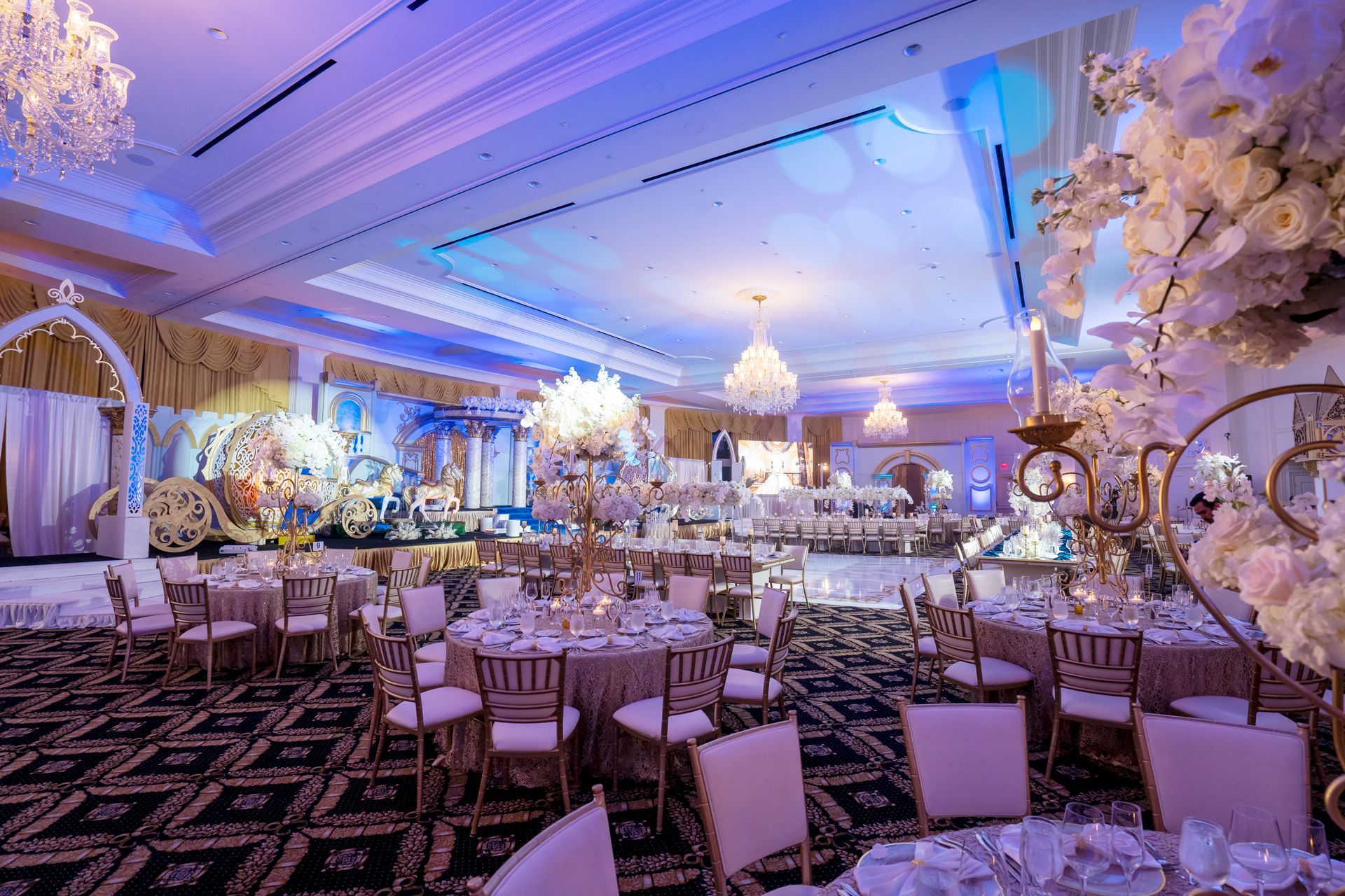 Lavishly decorated banquet hall with round tables, chandeliers, and blue-purple lighting