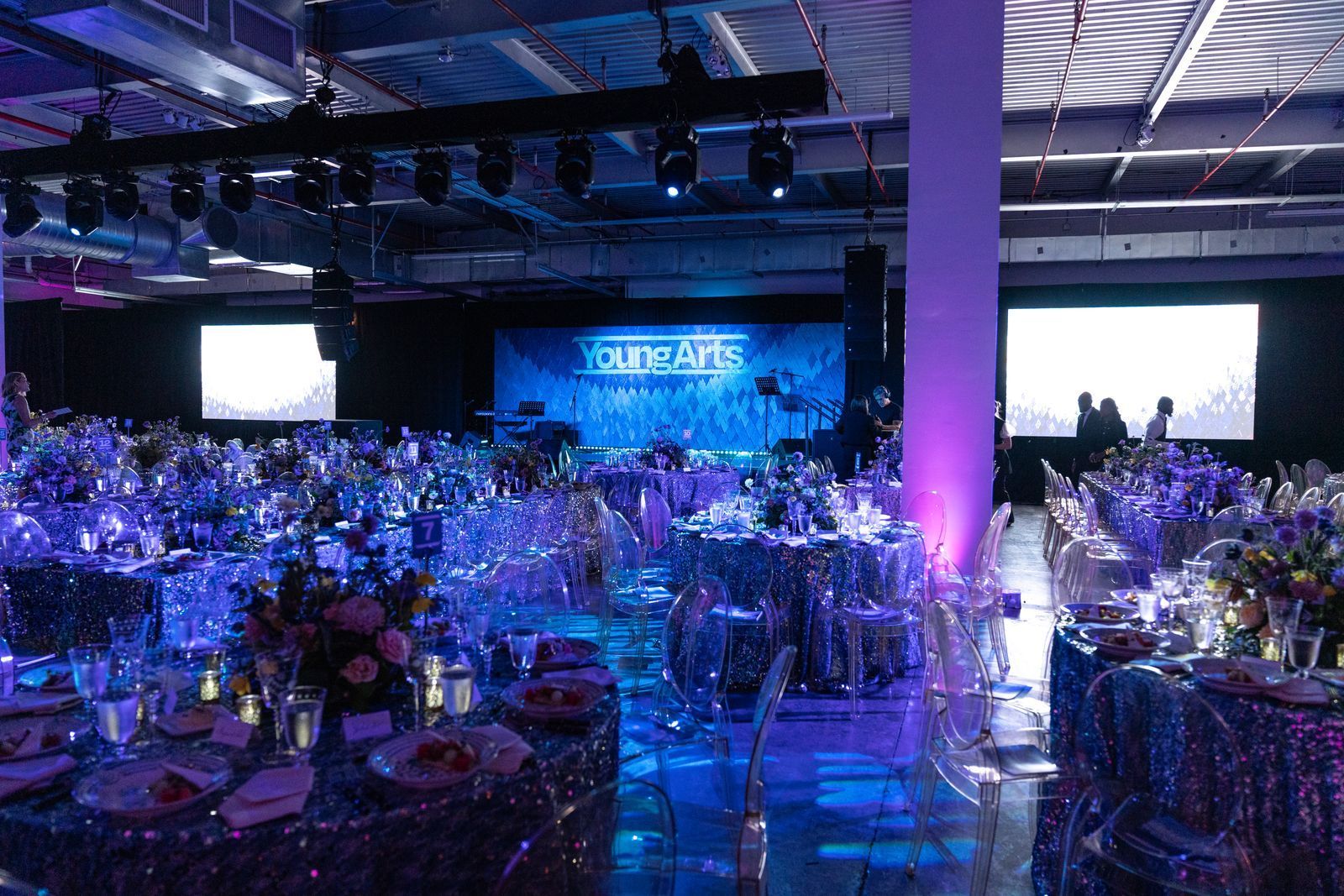 Lavishly decorated banquet hall with purple-blue lighting, round tables, and a stage with bright screens in the background