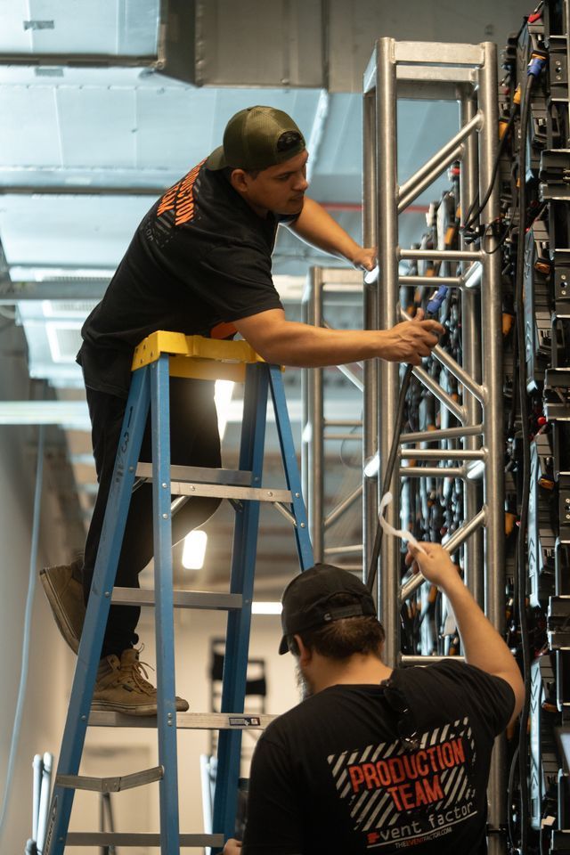 Workers assembling a metal rack on a ladder in an indoor industrial setting