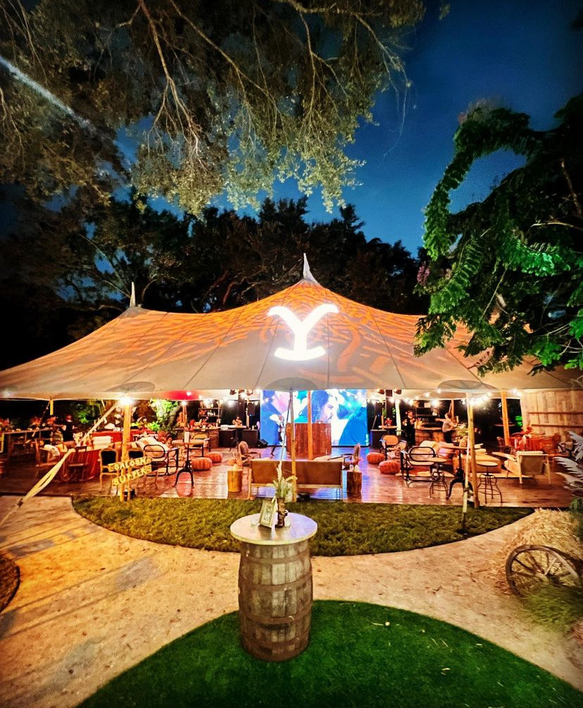 Outdoor tented event space at night with warm lights, bar seating, and a lit palm logo under trees