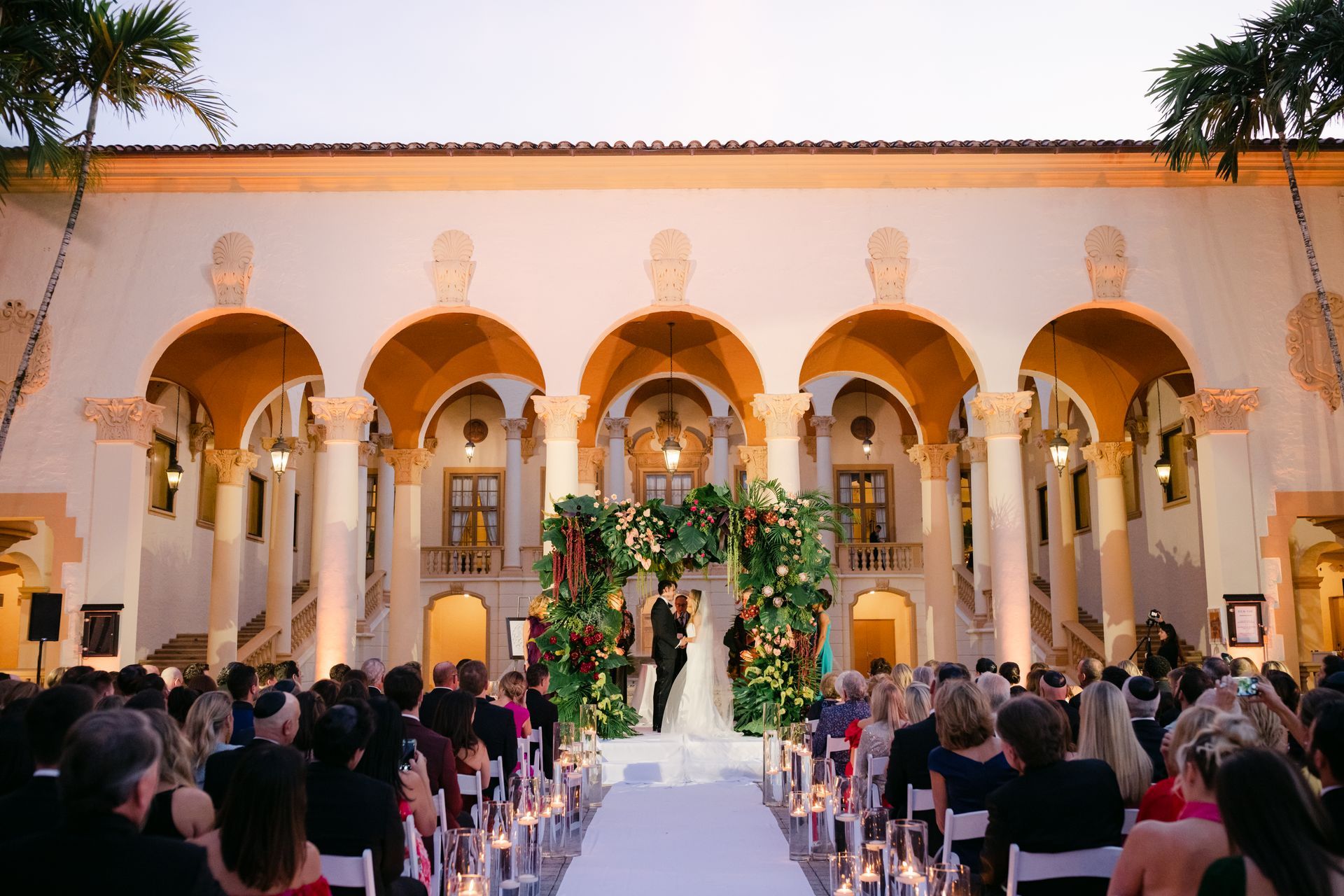 Wedding ceremony in a courtyard with arches, a floral altar, and guests seated along the aisle.