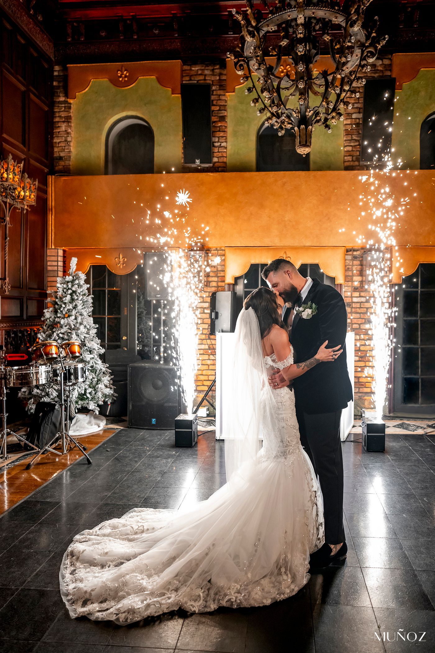Bride and groom kiss amid indoor fireworks in an ornate hall, bride in white gown with long train