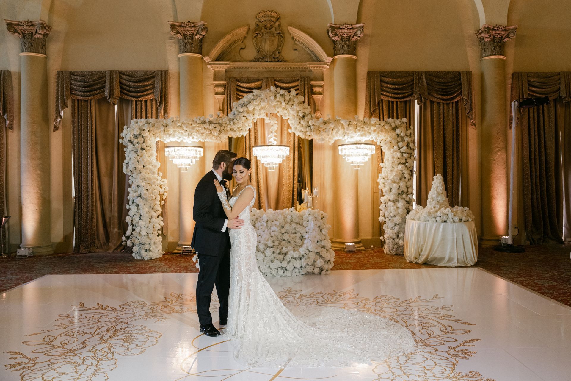 Bride and groom kissing in a lavish gold-decorated wedding hall under floral arches