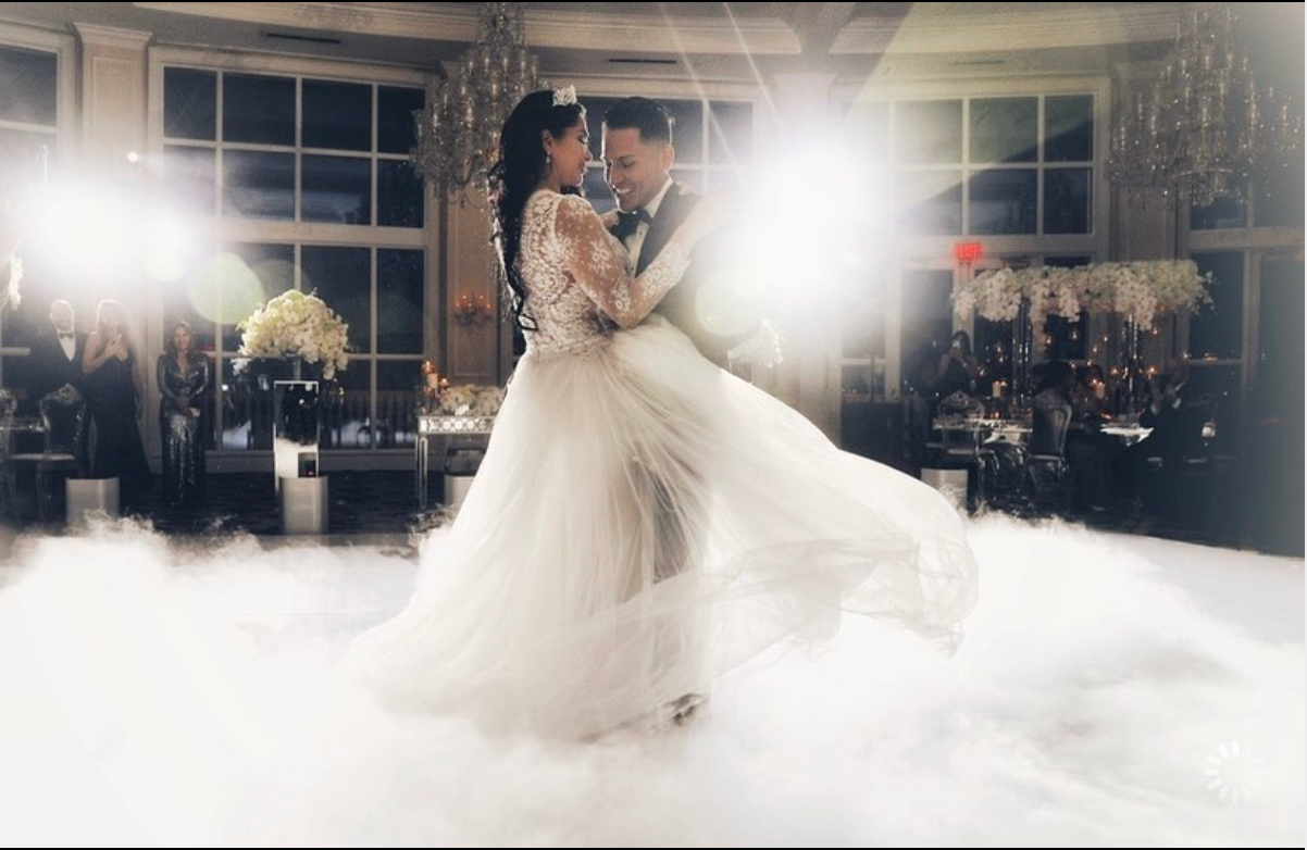 Bride and groom dancing in a white gown, with bright lights and clouds of fog in a ballroom