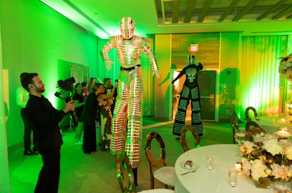 Costumed performers on stilts at a green-lit indoor event with guests and a decorated table