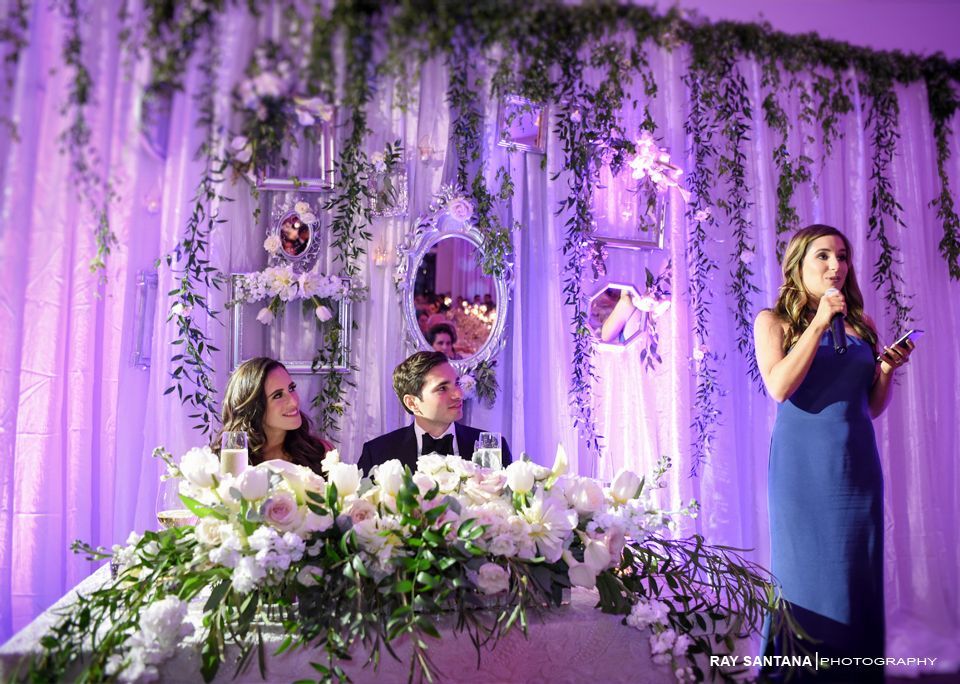 Woman speaking at a purple-lit wedding stage with seated guests and floral arrangements