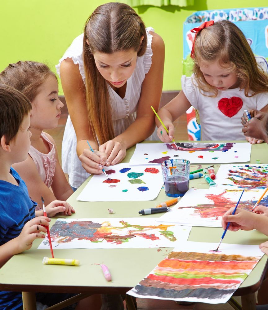 Teacher Helping Children Paint at a Table With Colorful Art Supplies and Paper Drawings — Bowen Childcare & Early Education in Bowen, QLD
