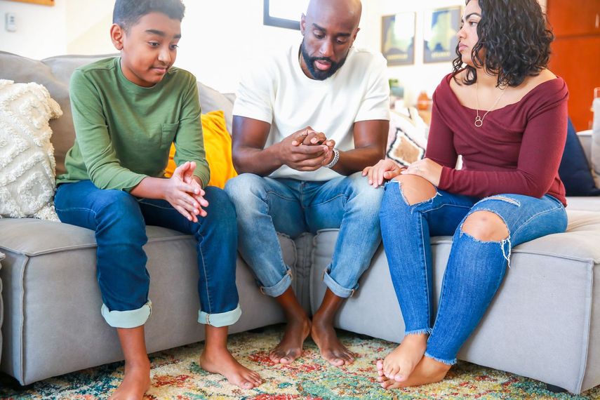 Three people sitting on a couch, looking at a phone in a bright living room.