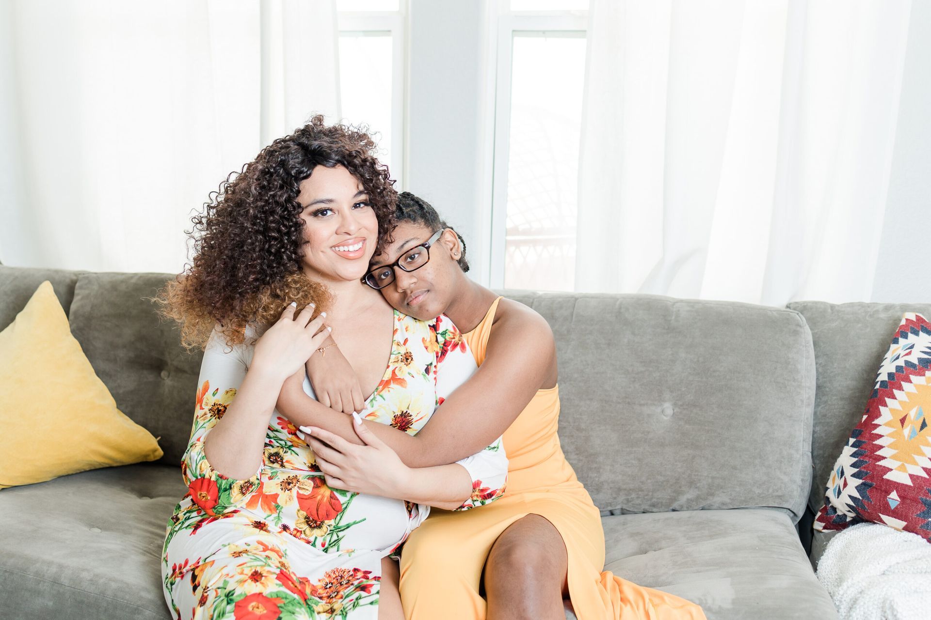 Two people embracing on a gray couch in a bright living room, one in a floral dress and one in a yellow outfit.