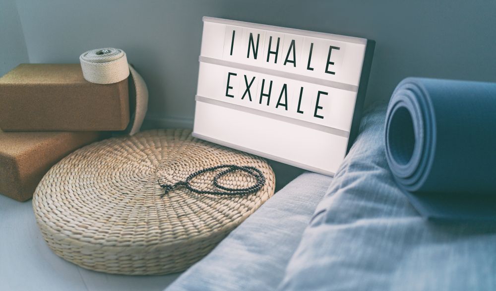 Yoga mat, cushion, and sign reading “Inhale Exhale” in a calm room with soft blue bedding.