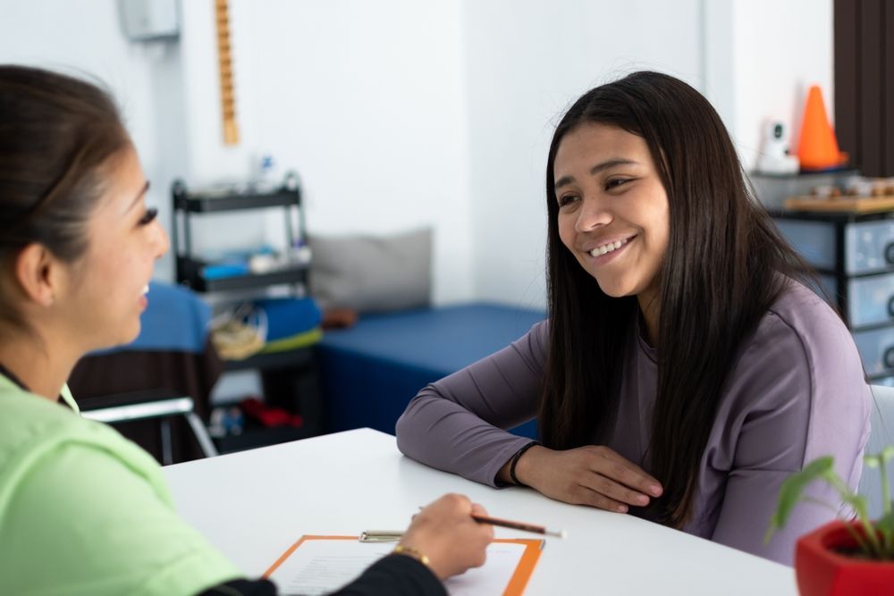 Two people talking at a table in a bright office, one smiling and holding a clipboard.