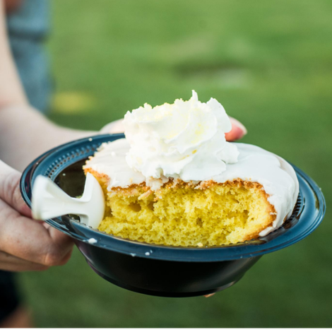 A slice of yellow cake with white frosting on a blue plate, held outdoors against a blurred green background