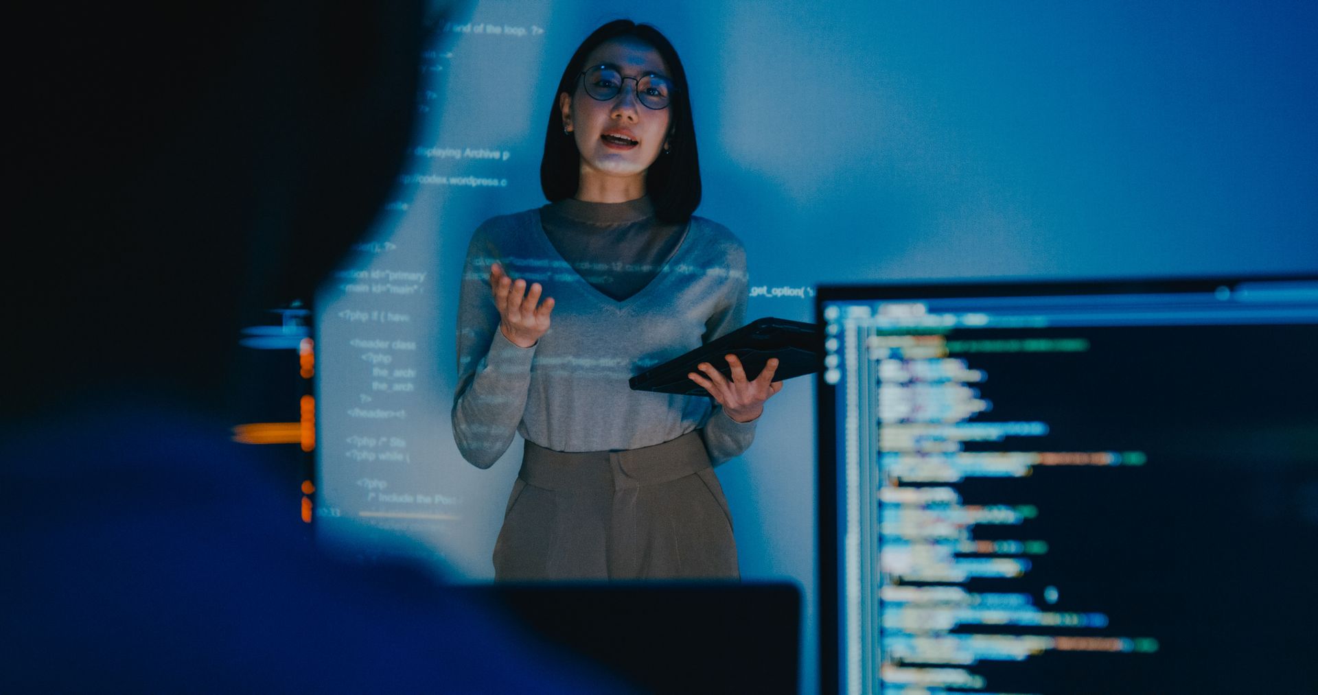 A woman is standing in front of a computer screen holding a tablet.