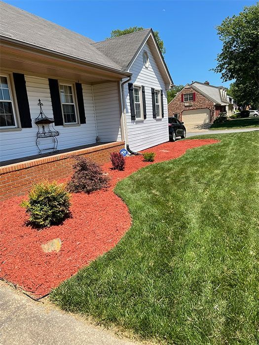 Red mulch borders house with green lawn, bushes, and a brick foundation under white siding.