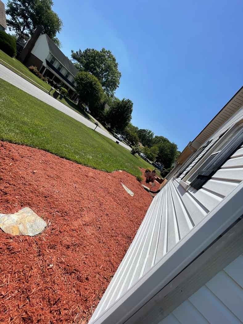 Red mulch bed next to a house with white siding, grassy hill, and a blue sky.