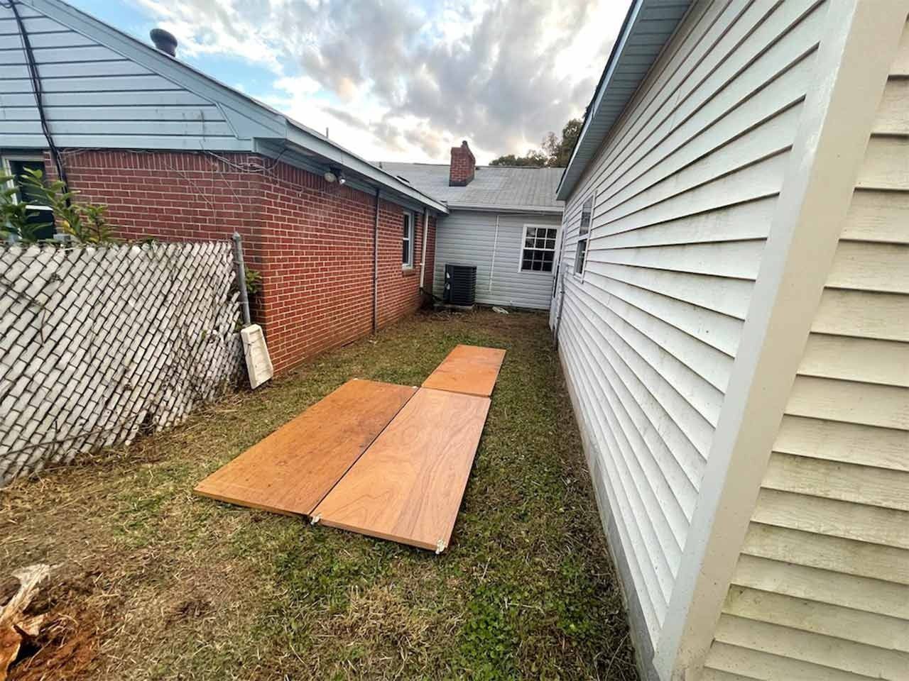 Narrow backyard between two buildings; wood planks on grass. Brick and white siding buildings.