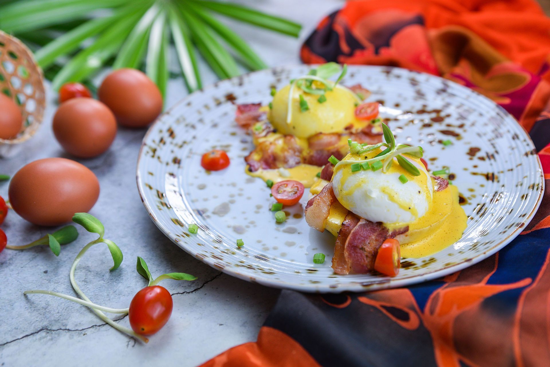 Eggs Benedict on a speckled plate, bacon, hollandaise, tomatoes, and chives, with eggs and a colorful scarf.