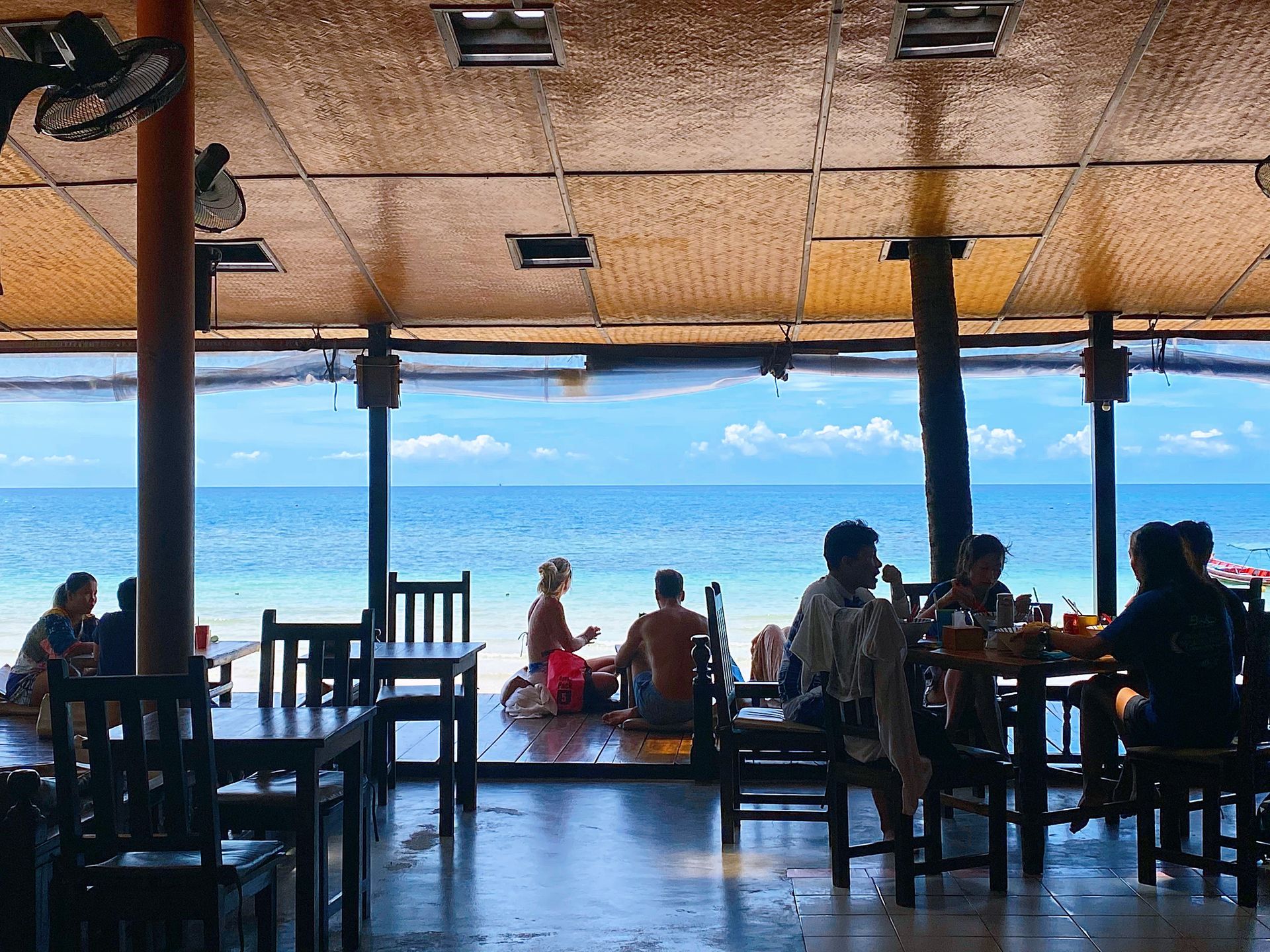 Beachside restaurant with tables and chairs, people eating, blue ocean visible.