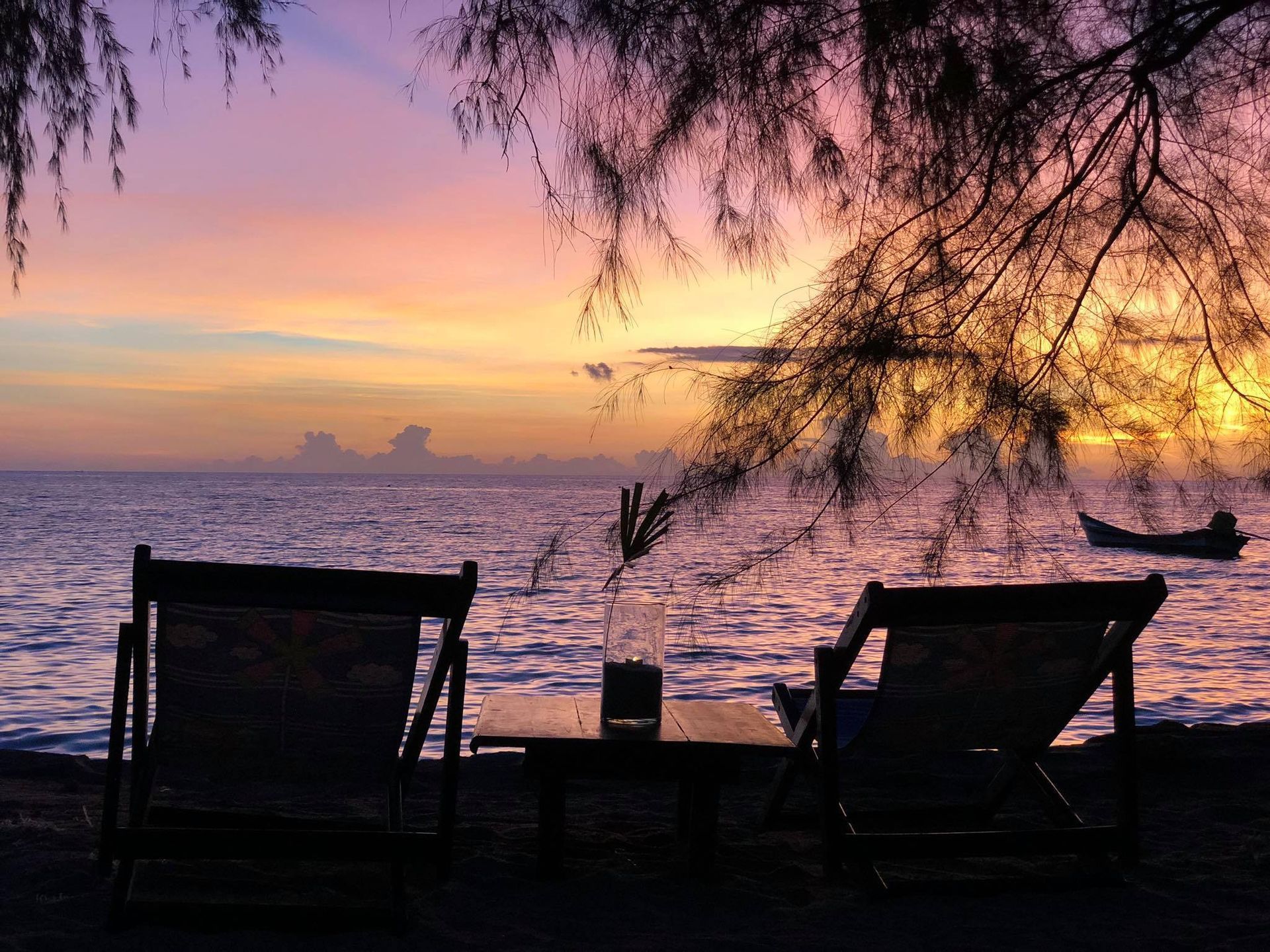 Beach scene at sunset: two chairs face the ocean. Purple and orange sky, small boat in the water, branches overhead.