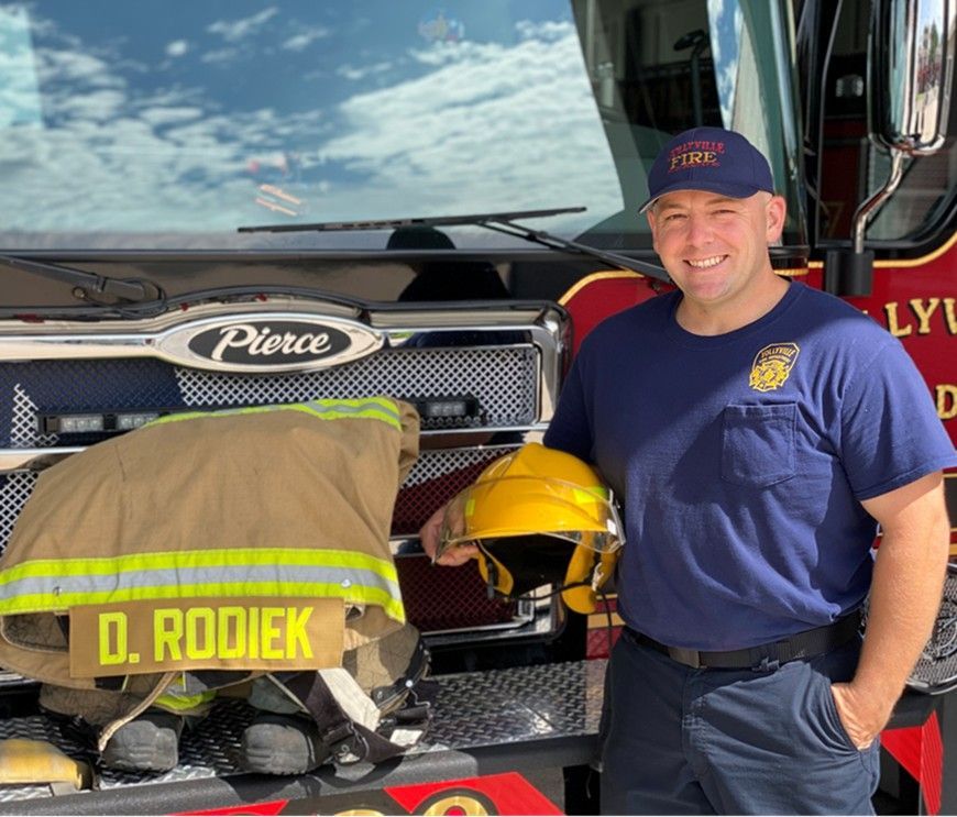 A picture of a firefighter standing in front of a fire engine with fire gear on the front bumper.