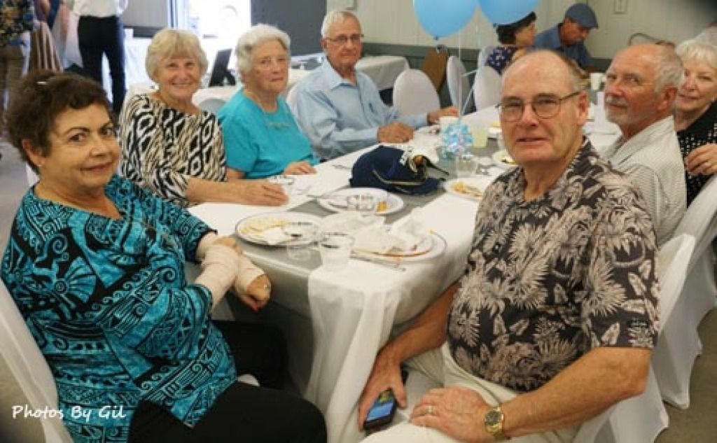 A group of eight older adults sit around a dining table, smiling at the camera.