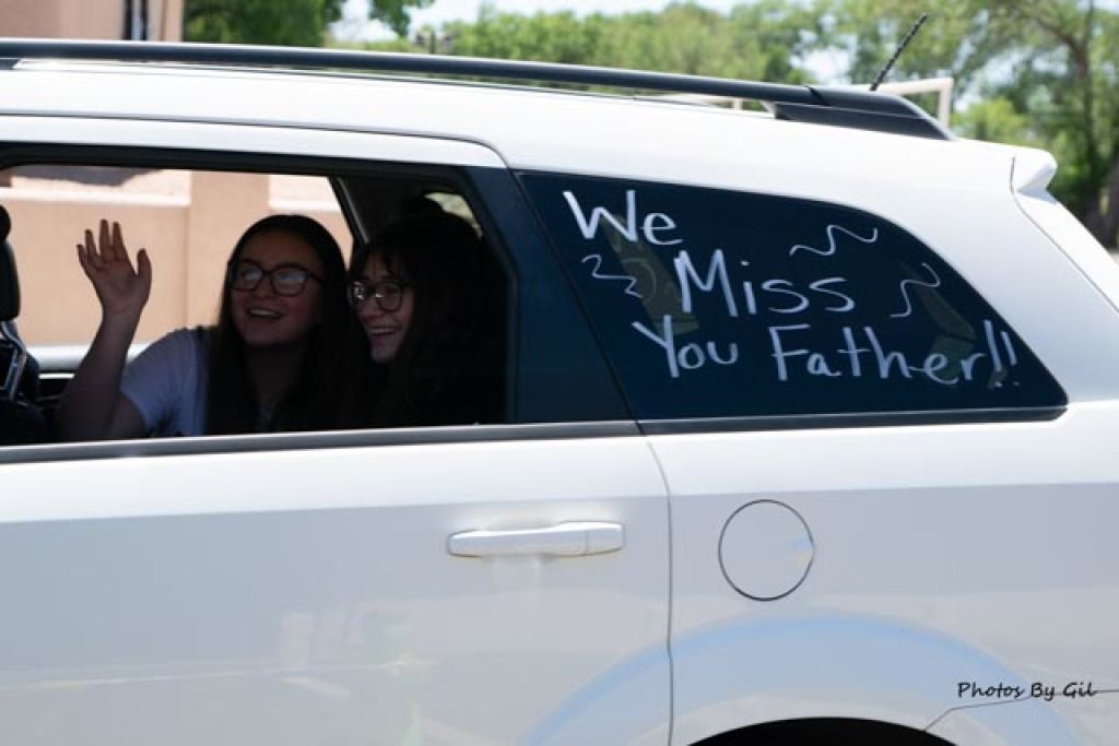 Two smiling individuals wave from the backseat of a white SUV. 
