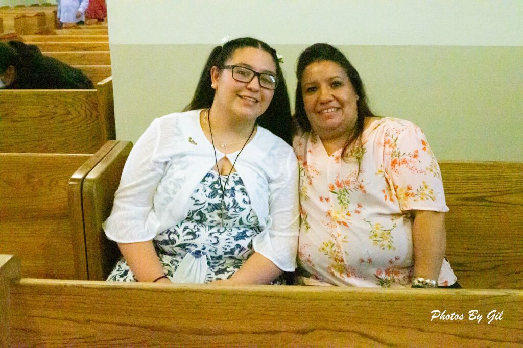 A woman and a girl sit closely on wooden pews, smiling warmly. 