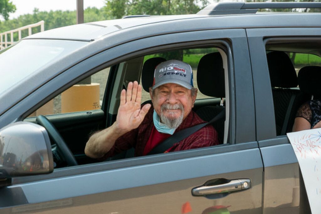 A smiling elderly man in a cap waves from the driver's seat of a parked SUV.