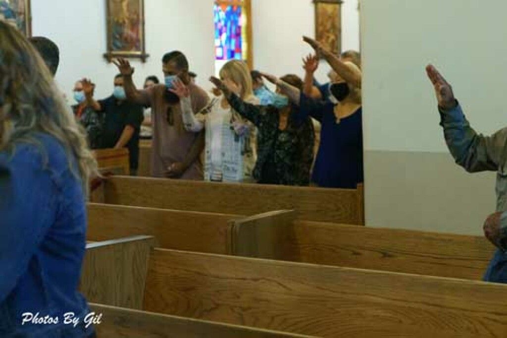 People in a church pew raise one hand, possibly in prayer or blessing. 