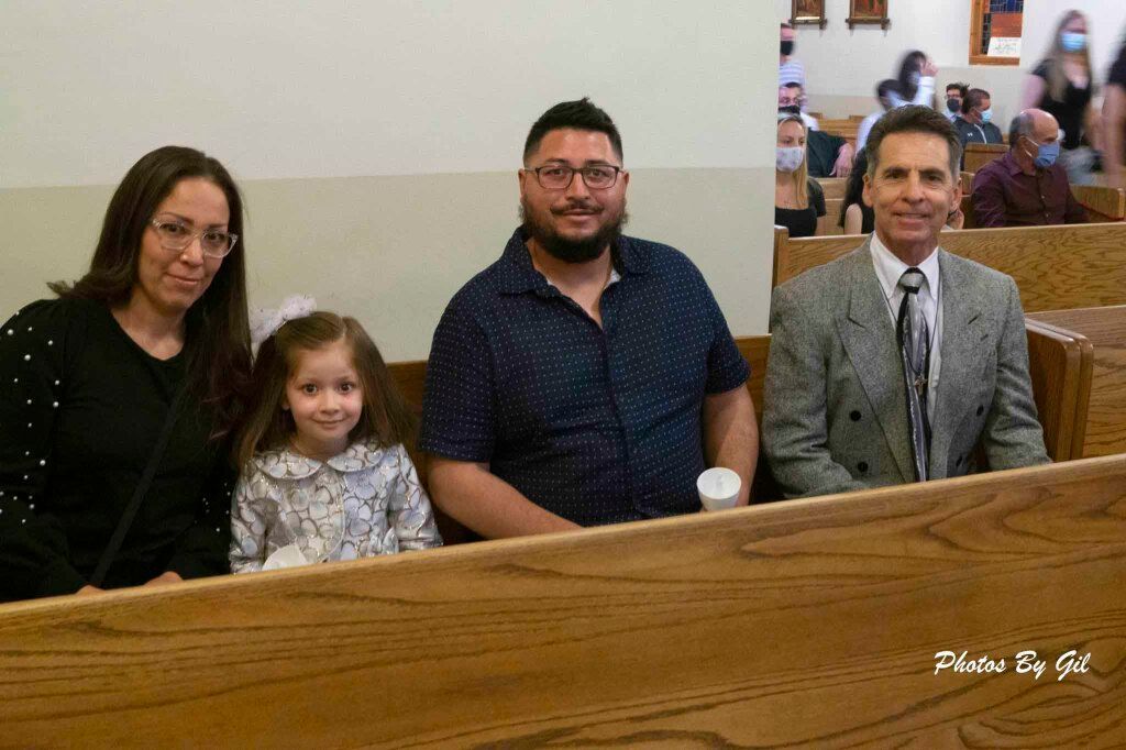 A family of four sits in a church pew, a woman, child, man, and older man smile toward the camera. 