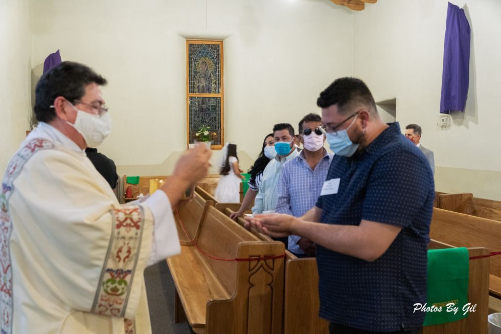 Priest in white robe, masked, gives communion to a young man in a church. 