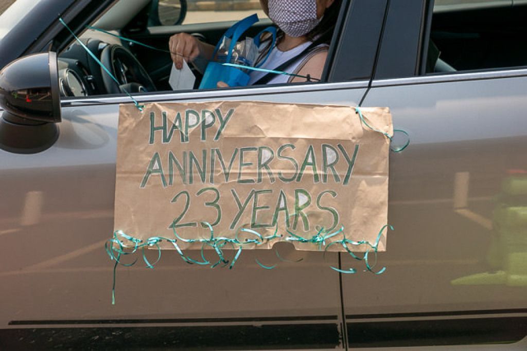 A masked person is in a car, holding a Happy Anniversary sign.