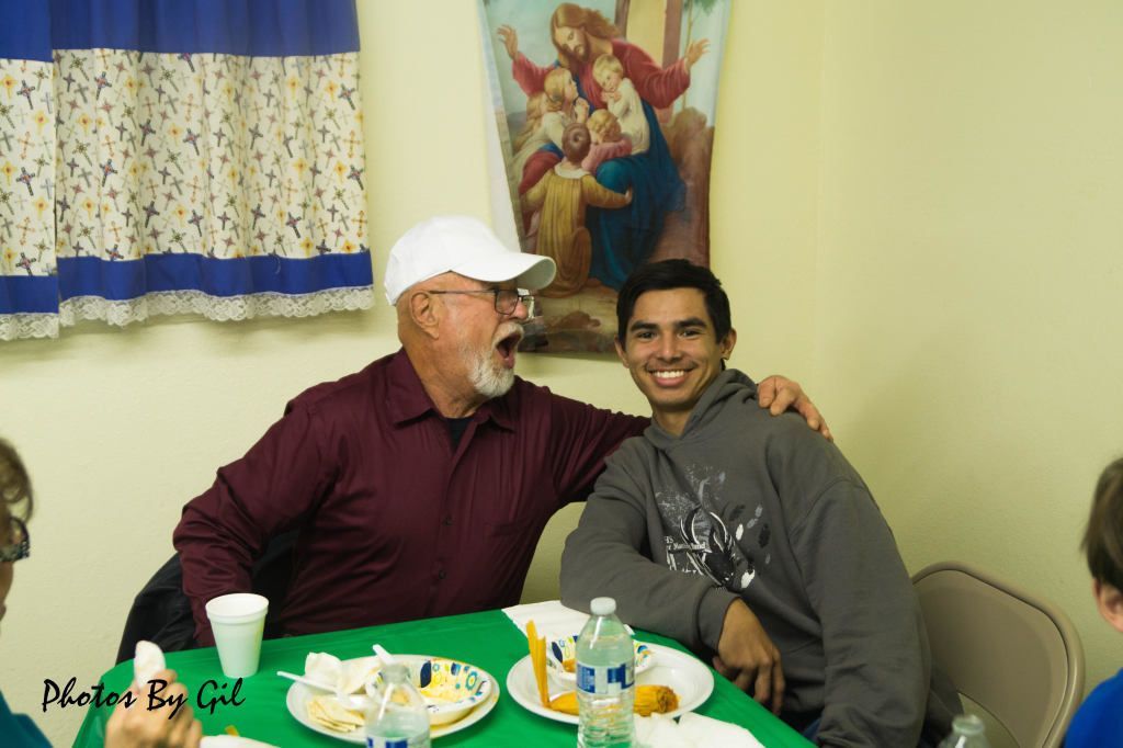 Two smiling men sit at a table with food and drinks. 