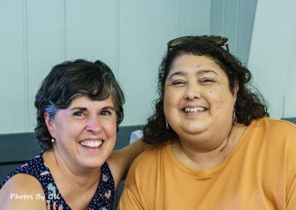 Two women are smiling warmly at the camera, seated indoors against a light blue wall.