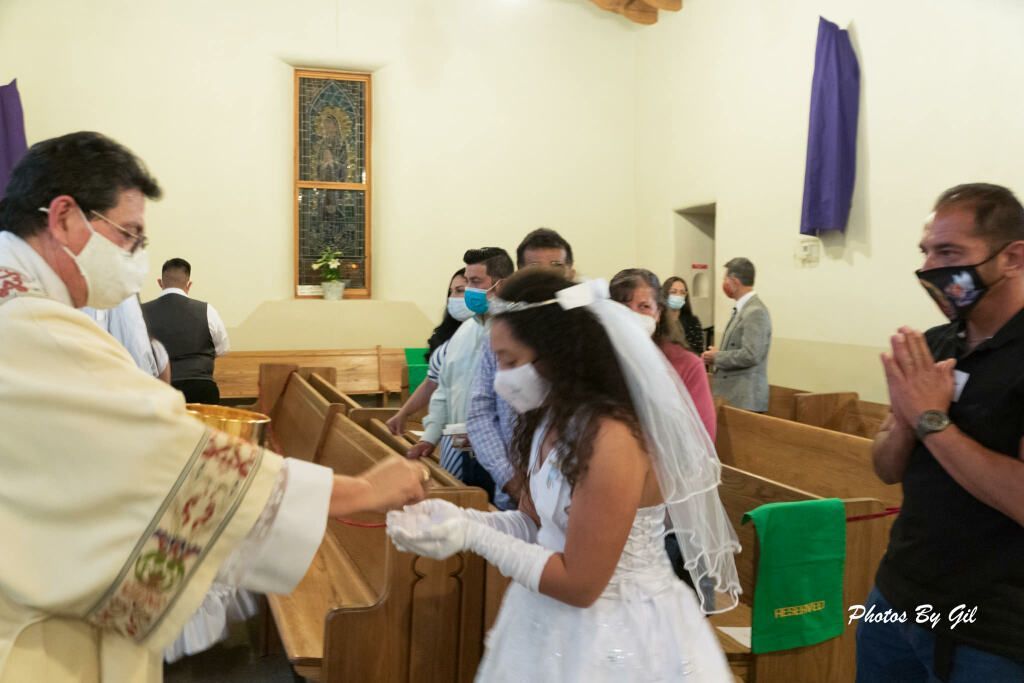 A priest wearing a mask gives communion to a young girl in a white dress and veil during a church ceremony. 