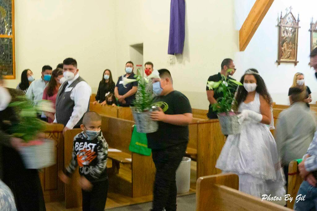 A group of people, including a child in a white dress and tiara, carry potted plants down a church aisle. 