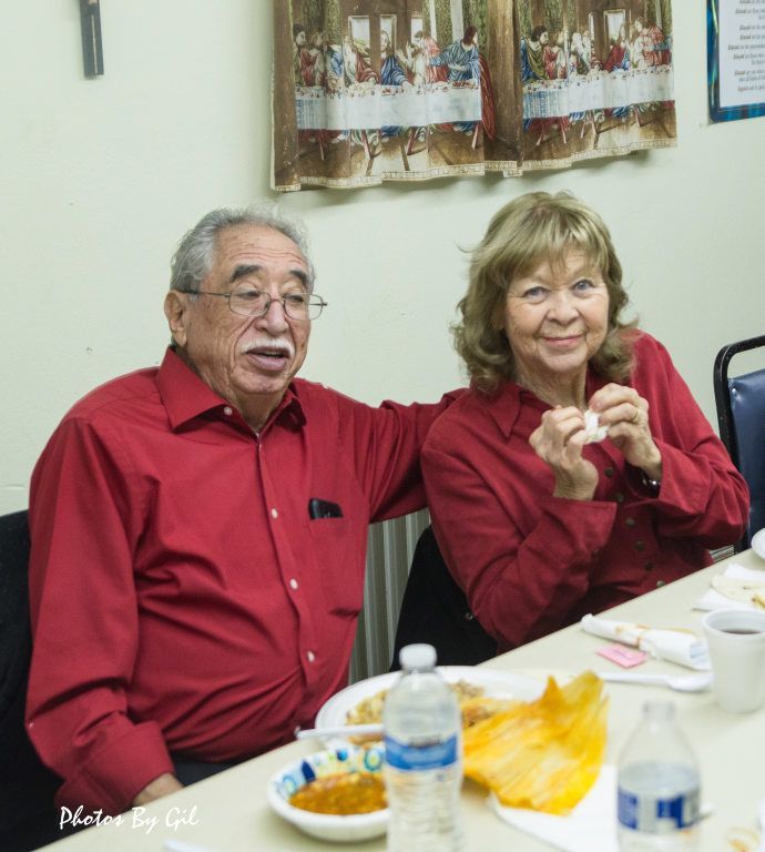 Elderly couple in red shirts sits at a dining table, smiling.