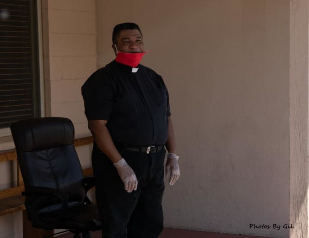 A person in a clerical shirt with a red mask and gloves stands outside near a wall and an office chair. 
