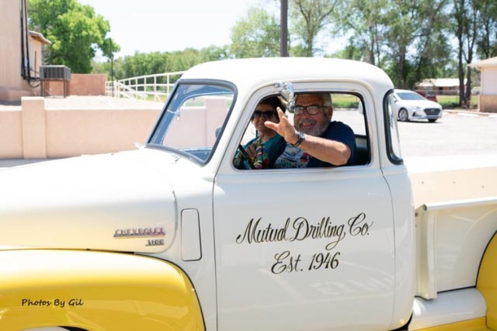 A man and woman wave from a vintage white and yellow Chevrolet truck.