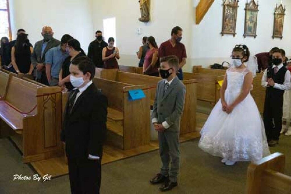Children in formal attire stand in a church aisle, wearing face masks, appearing solemn.