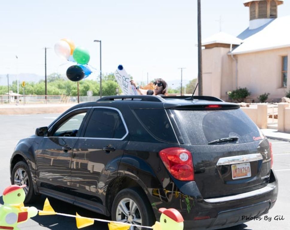 A person joyfully holds a graduation sign through a black SUV's sunroof.