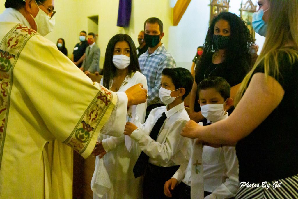 A priest conducts a Catholic ceremony, blessing masked children in white clothing and ties.