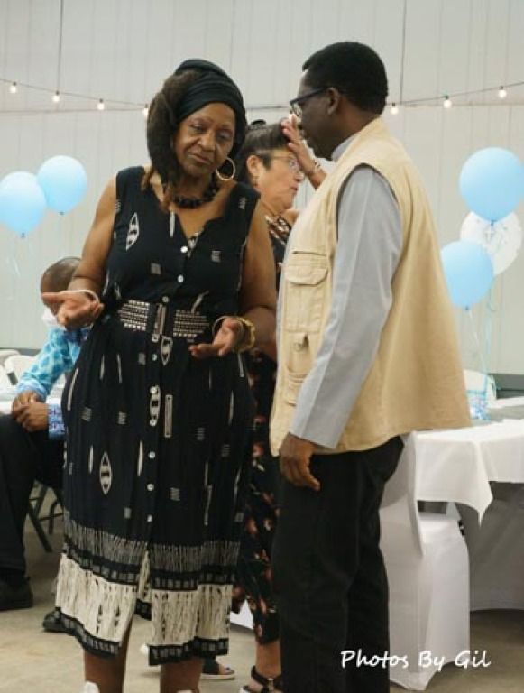 A woman in a patterned black dress speaks animatedly to a man in a beige vest at an indoor event.