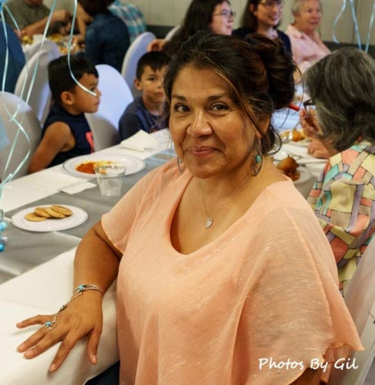 A woman in a peach blouse smiles at a dining table, surrounded by people at a gathering. 
