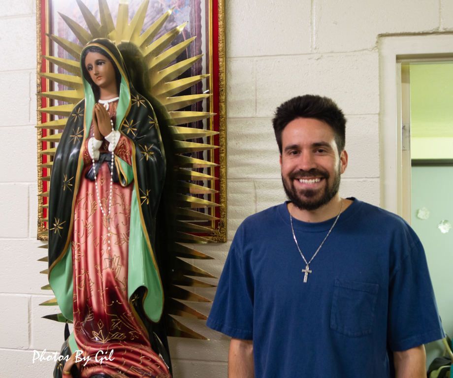 A smiling man in a blue shirt stands beside a statue of Our Lady of Guadalupe.