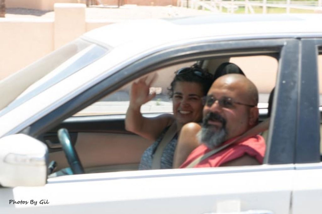 A woman waves cheerfully while seated in the passenger seat of a car, next to a man driving. 