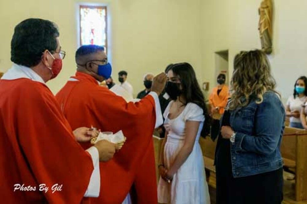A priest in red robes blesses a young girl in a white dress during a church ceremony.
