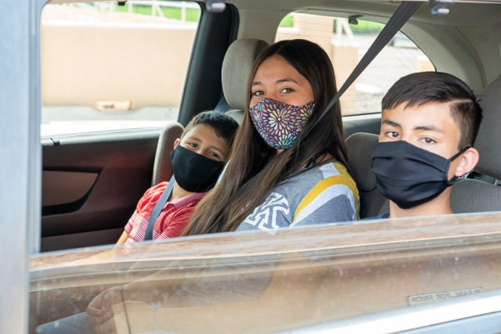 A woman and two children in face masks sit in a car's backseat, looking out the window. 