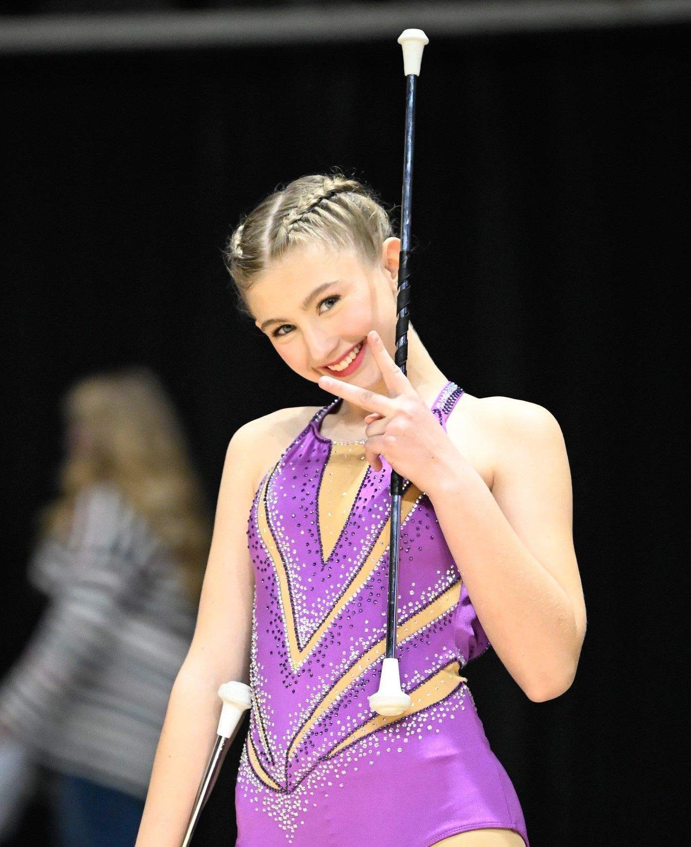 Girl in purple leotard smiles, holds baton, gives peace sign. Dark background.